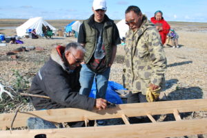 One man sitting behind a sled teaching two men who stand above him how to build the frame